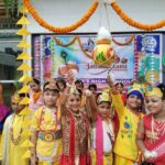 Group of smiling indian schoolchildren in colorful traditional costumes at a festive ceremony with marigold garlands and a hanging decorative pot in the foreground